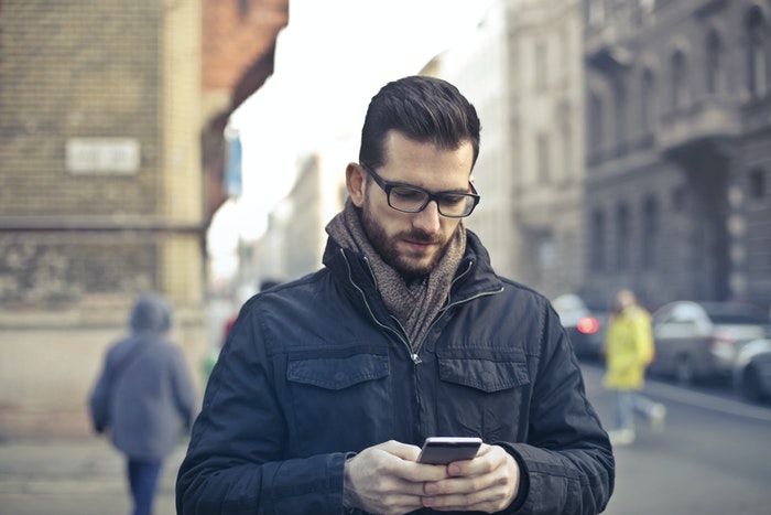 Man Wearing Black Zip Jacket Holding Smartphone Surrounded by Grey Concrete Buildings