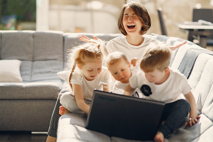 Stressed woman asking for help while sitting with small children playing on laptop together on sofa in living room